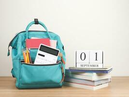 Front view  of green backpack with school supplies, stack of books and wooden calendar September 01 on wooden table and white  background with copy space. photo