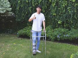 Asian young man wearing aircast boot walking in the garden with orthopedic walker. showing thumb up and looking at camera. photo