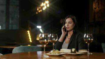 sonriente bonito joven mujer hablando en móvil teléfono sentado a mesa con rojo vino en moderno restaurante con oscuro interior. atractivo contento hembra teniendo conversacion en teléfono inteligente en bar. video