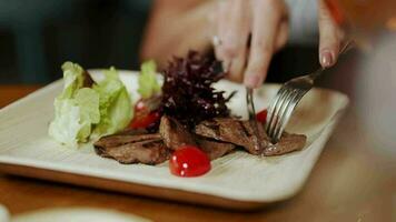 gegrillt Steak mit Tomaten und Grün Grüner Salat Salat mit braten Zwiebel und Chili Pfeffer auf Gericht Über hölzern Tisch. ein Mädchen schneidet ein Steak auf ein Teller mit ein Gabel und ein Messer video