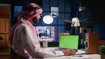 Middle Eastern man scrolling on green screen laptop, checking social media posts. Self employed man browsing online websites from mockup device in apartment office with TV as background noise video