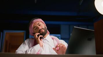 Muslim worker answering phone call while remotely working from cozy apartment office. Middle Eastern teleworker talking with colleague using smartphone, discussing paperwork and tasks needing to be video