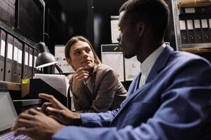 Investigation team working overhours at fraude case, analyzing new evidence in arhive room. Diverse private detectives reading police expertise, trying to catch suspect. Criminology concept photo