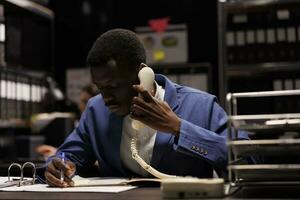 africano americano gerente hablando a teléfono fijo teléfono, que se discute administración investigación con remoto contable en almacenamiento habitación. empresario trabajando tarde a noche a contabilidad reporte foto