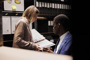 Diverse depository employees analyzing bureaucracy record, checking administrative documents late at night in storage room. Bookkeepers discussing accountancy report, discovering management files photo