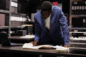 African american manager working at accountancy report, analyzing administrative files in company repository. Businessman checking bureaucracy record, searching for management research photo