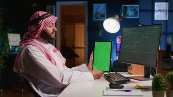 Muslim worker in apartment office holds chroma key tablet, crosschecking data on it with business report graphs on computer. Smiling entrepreneur with mockup digital device in hand performs job tasks video