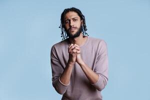 Arab man with clenched hands looking at camera with pleading facial expression. Person praying with folded arms gesture while asking for forgiveness with hopeful emotions photo