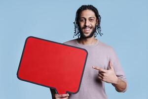 sonriente árabe hombre señalando con dedo a rojo habla burbuja con Copiar espacio retrato. alegre contento persona participación vacío bandera y publicidad producto mientras mirando a cámara foto