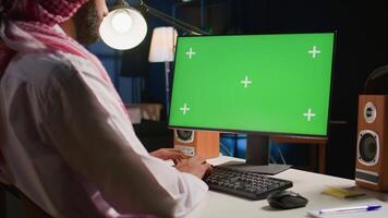 Muslim worker looking at chroma key computer screen while working from home. Arab employee typing on keyboard, sending emails on green screen device in apartment office with TV as background noise video