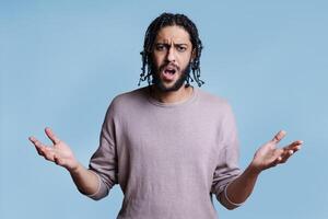 Angry arab man with open arms and confused facial expression portrait. Young attractive dissatisfied person with negative emotion shouting and looking at camera while posing for studio shot photo