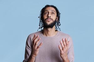 Arab man praying with open hands while looking upwards. Religious young person pleading for god blessing and begging for forgiveness while holding palms up studio shot on blue photo