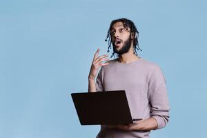 Puzzled arab man thinking about software project while holding laptop and looking upwards in doubt. Pensive person with thoughtful facial expression while coding on portable computer photo