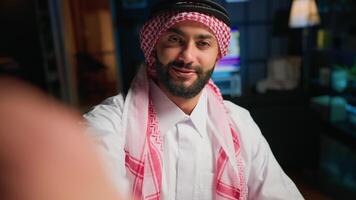 POV of cheerful Muslim content creator dressed in traditional clothing filming himself. Smiling Arab man with thobe and headscarf waving towards camera, doing online vlogging video