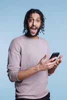 Excited arab man using smartphone and looking at camera with happy facial expression. Cheerful person in casual clothes getting online message with good news on mobile phone portrait photo