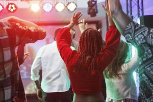 People dancing and raising hands while attending dj party in nightclub. Clubbers having fan and jumping on crowded dancefloor at discotheque in club illuminated with vibrant lights photo