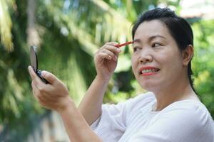 Asian middle aged woman is making up, drawing her eyebrows, looking at portable mirror, outdoor background. Concept, women don't stop beauty, using cosmetics. Woman lifestyle, make self looks good. photo