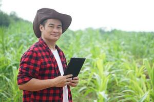 Handsome Asian man farmer is in garden, wears hat, red plaid shirt, holds  smart tablet. Feel confident, point finger, proud in crops. Concept, Agriculture occupation, inspect, survey and research. photo