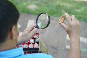 cerca arriba hombre sostener aumentador vaso a inspeccionar detalle y modelo minúsculo de amuleto desde recopilación. concepto, Fé creencia en santo y suerte para budistas colectar amuleto como aficiones, intercambiar o comercio. foto