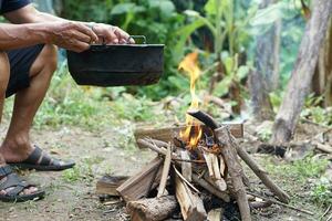 cerca arriba hombre sostiene antiguo negro maceta a cocinar en hoguera. concepto, Cocinando exterior, cocina en bosque. supervivencia vida habilidad para cámping o senderismo. rural tradicional estilo de vida. Encendiendo fuego con madera. foto