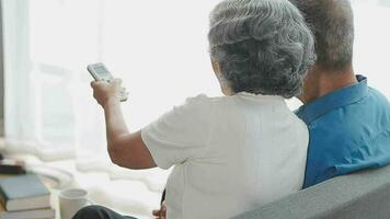 Happy senior couple using remote control to change channel. Seniors watch TV for entertainment while man using remote control. Wife and old man sitting on couch in living room with television show. video