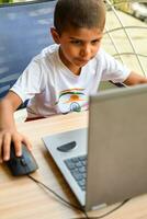 pequeño chico sentado a mesa utilizando ordenador portátil para en línea clase en grado 1, niño estudiando en ordenador portátil desde hogar para distancia aprendizaje en línea educación, colegio chico niños estilo de vida concepto foto