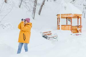 contento niña fotógrafo en un amarillo chaqueta toma imágenes de invierno en un Nevado parque foto