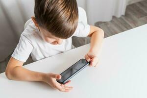 A little boy in a white T-shirt plays games on the phone at home. A happy child looks at his smartphone. photo