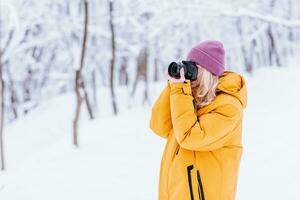 contento niña fotógrafo en un amarillo chaqueta toma imágenes de invierno en un Nevado parque foto