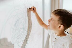 A cheerful boy with a pencil stands near the wall that he painted. A child is engaged in creativity at home. photo