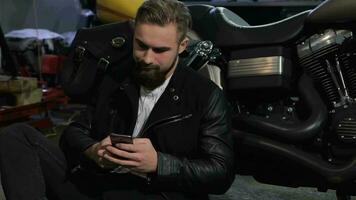 Male biker uses his phone near the chopper at the motorcycle workshop video