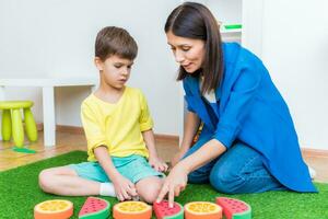 A woman speech therapist deals with a boy and performs an exercise to correct the speech apparatus by playing on the floor photo