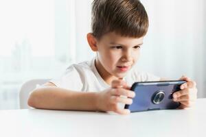 A little boy in a white T-shirt plays games on the phone at home. A happy child looks at his smartphone. photo