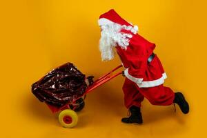 Papa Noel claus es corriendo con un saco lleno de regalos en un metal carretilla en naranja antecedentes foto
