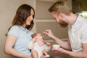 Parents feed their daughter fruit puree together in the kitchen photo