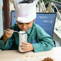 Cute Indian chef boy preparing sundae dish as a part of non fire cooking which includes vanilla ice cream, brownie, coco powder, freshly chopped fruits and strawberry syrup. Little kid preparing food photo