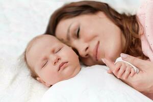 Young beautiful woman holding small hand of her sleeping baby daughter in bedroom photo
