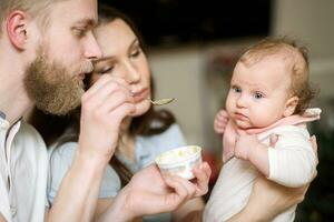 padres alimentar su hija Fruta puré juntos en el cocina foto