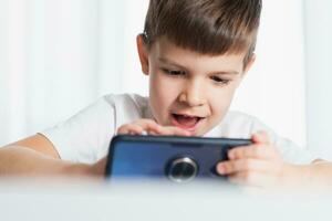 A little boy in a white T-shirt plays games on the phone at home. A happy child looks at his smartphone. photo