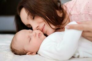 joven mujer cariñosamente mira a su dormido recién nacido hija en el dormitorio foto