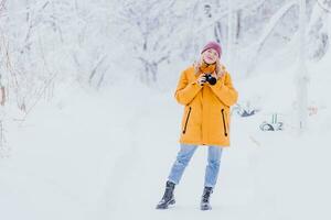 Happy girl photographer in a yellow jacket takes pictures of winter in a snowy park photo