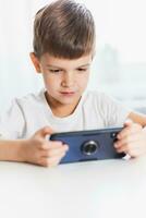 A little boy in a white T-shirt plays games on the phone at home. A happy child looks at his smartphone. photo