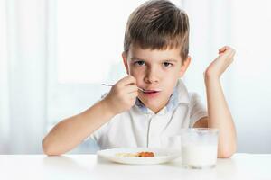 Cute child in white shirt eats cottage cheese pie and drinks milk for breakfast at home photo