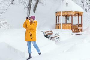 Happy girl photographer in a yellow jacket takes pictures of winter in a snowy park photo