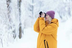 Happy girl photographer in a yellow jacket takes pictures of winter in a snowy park photo