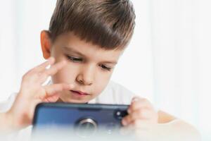 un pequeño chico en un blanco camiseta obras de teatro juegos en el teléfono a hogar. un contento niño mira a su teléfono inteligente foto