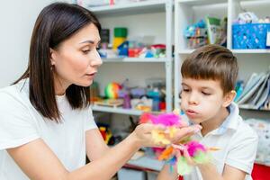 Woman speech therapist training boy to blow correctly using colored feathers exercise photo