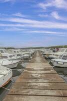 View along a wooden jetty in a harbor with docked ships in front of blue skies photo