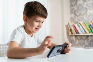 A cheerful child in a white T-shirt plays games on the phone at home. Happy boy looks at his smartphone. photo