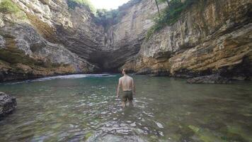 el joven hombre camina mediante el agua en el playa cueva con un fascinante vista. video